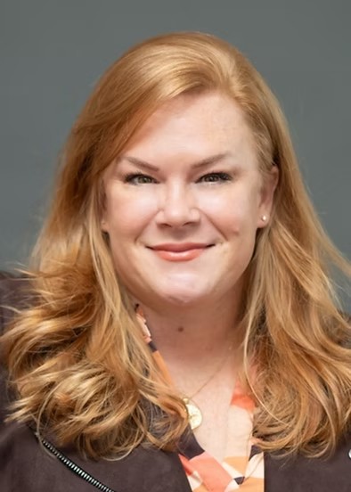 Woman with reddish-blonde hair in orange and brown shirt and brown jacket